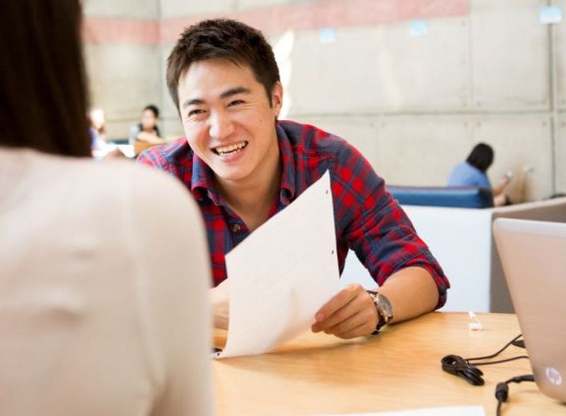 A man sits at a desk with a piece of paper smiling while talking to a woman across from him